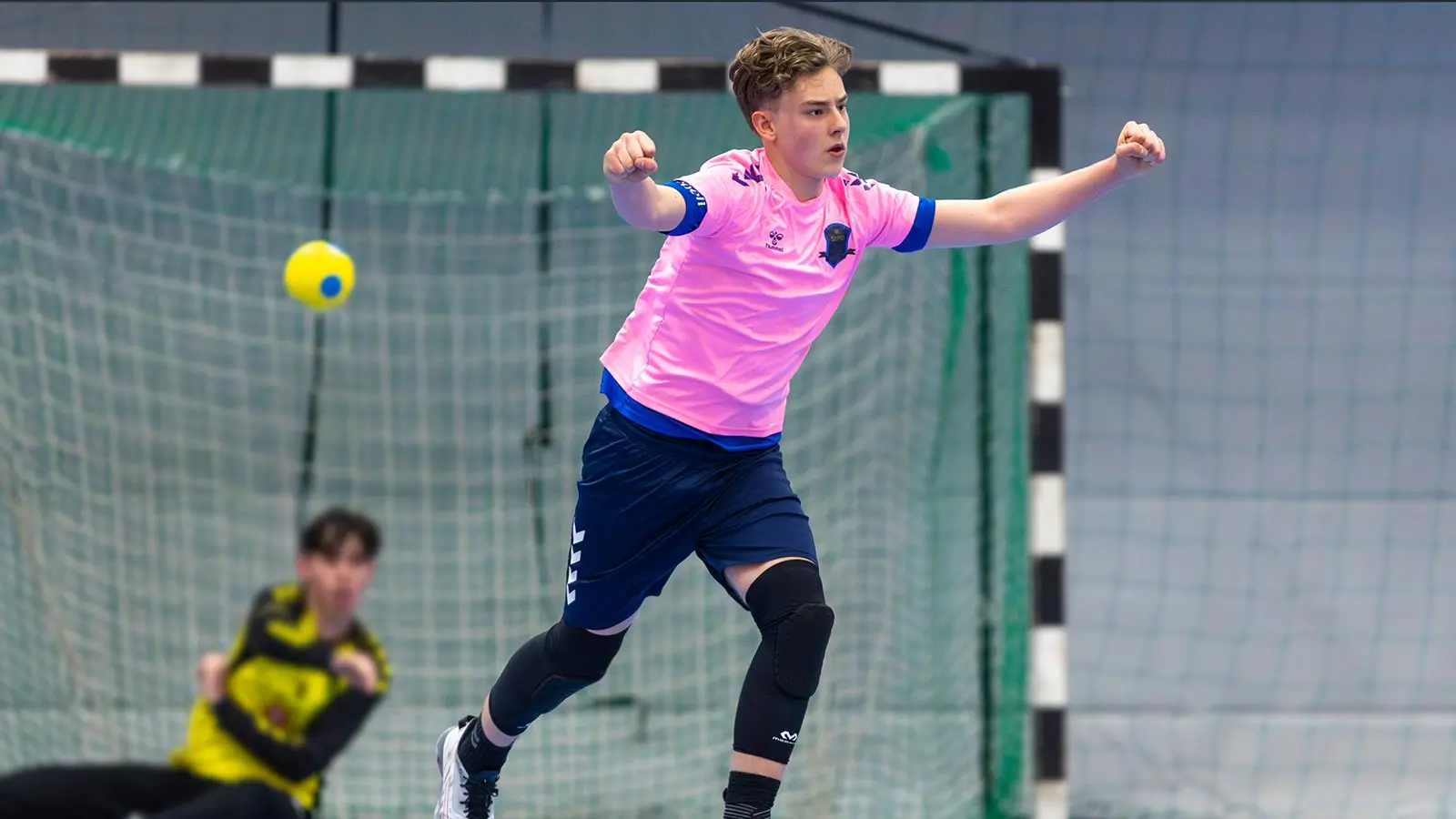 En handbollsspelare, kille, springer med armarna uppåtsträckta i en segergest. I bakgrunden syns suddigt en målvakt sittandes på golvet.